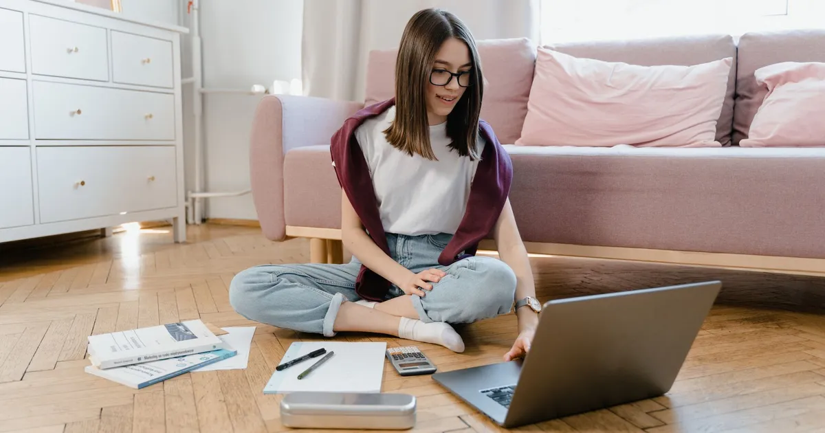 Teen girl studying online at home, sitting on the floor with a laptop and documents.