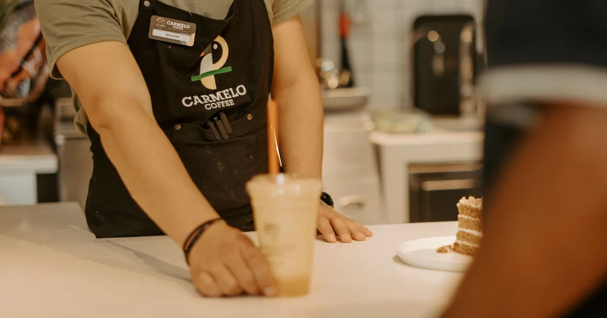A barista wearing an apron serves an iced coffee to a customer inside a coffee shop.