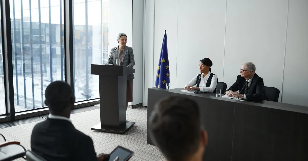 Professional conference setting with a speaker at a podium and attentive audience in a modern meeting room.