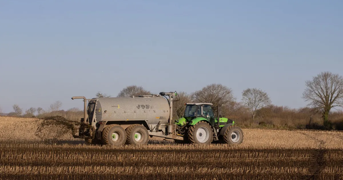 A green tractor spreading fertilizer on a brown, freshly plowed farmland under a clear sky.