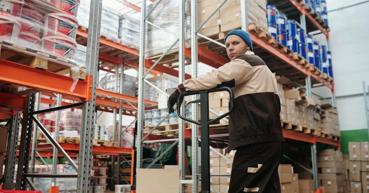 Warehouse worker operating a pallet jack among stacked boxes and metal shelves in an industrial setting.