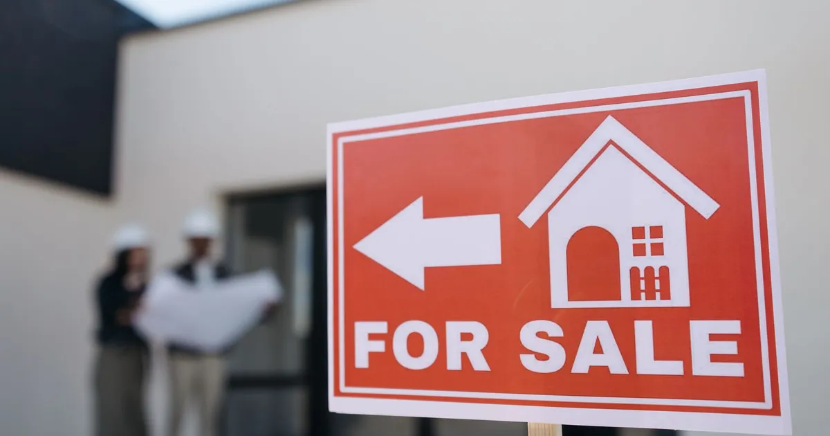 A real estate sign indicates a property for sale as two agents in hard hats discuss building plans outdoors.