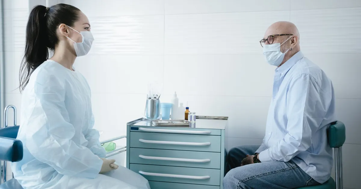 Medical professional consults a senior patient, both wearing masks in a clinic setting.
