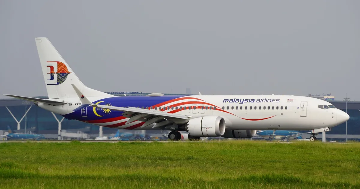 Malaysia Airlines aircraft on runway with terminal in background on a clear day.