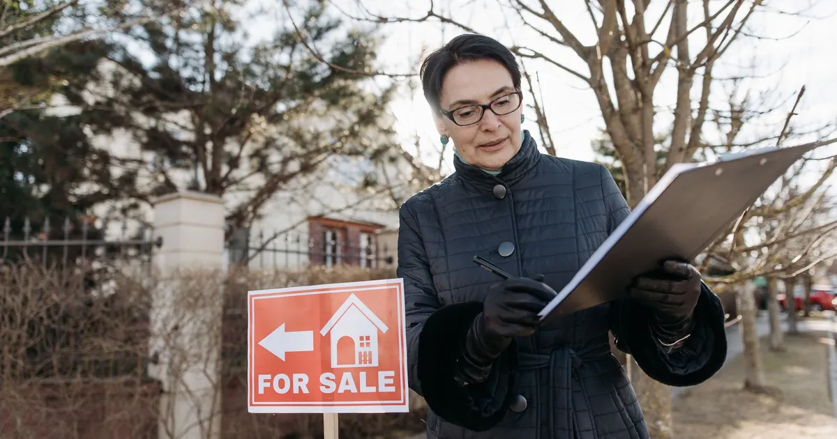 A real estate agent reviews a clipboard next to a 'For Sale' sign outside a property on a clear day.
