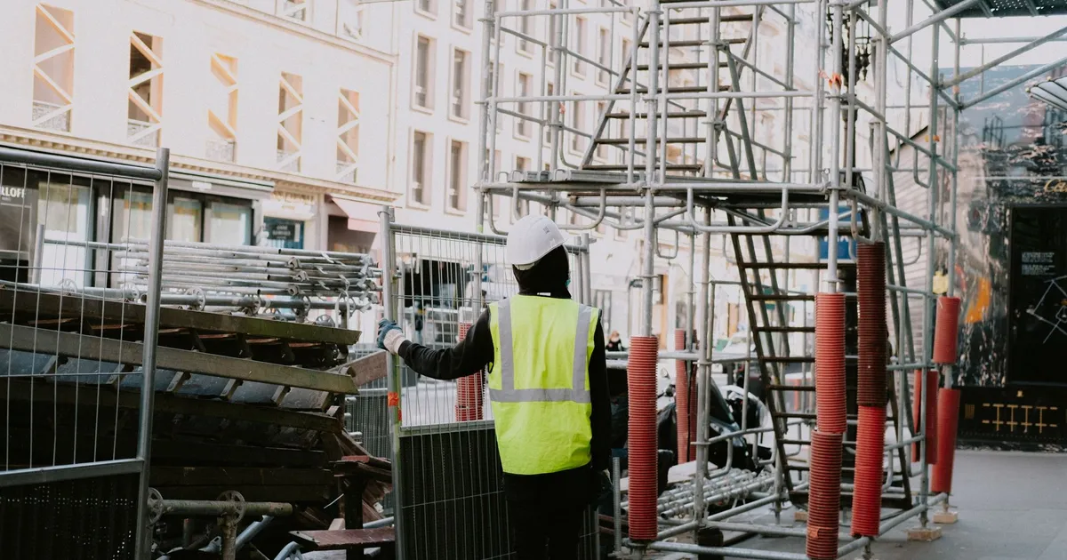 A construction worker onsite in a city wearing safety gear and examining scaffolding.