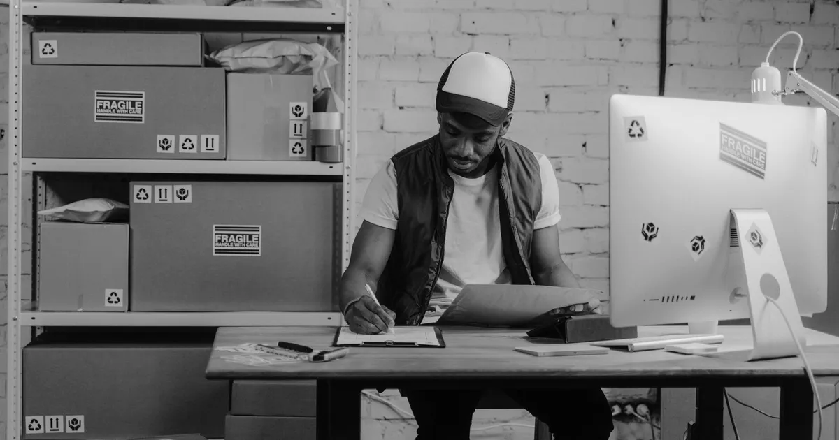 Warehouse worker organizing shipments at a desk, surrounded by packages and a computer, illustrating logistics work.