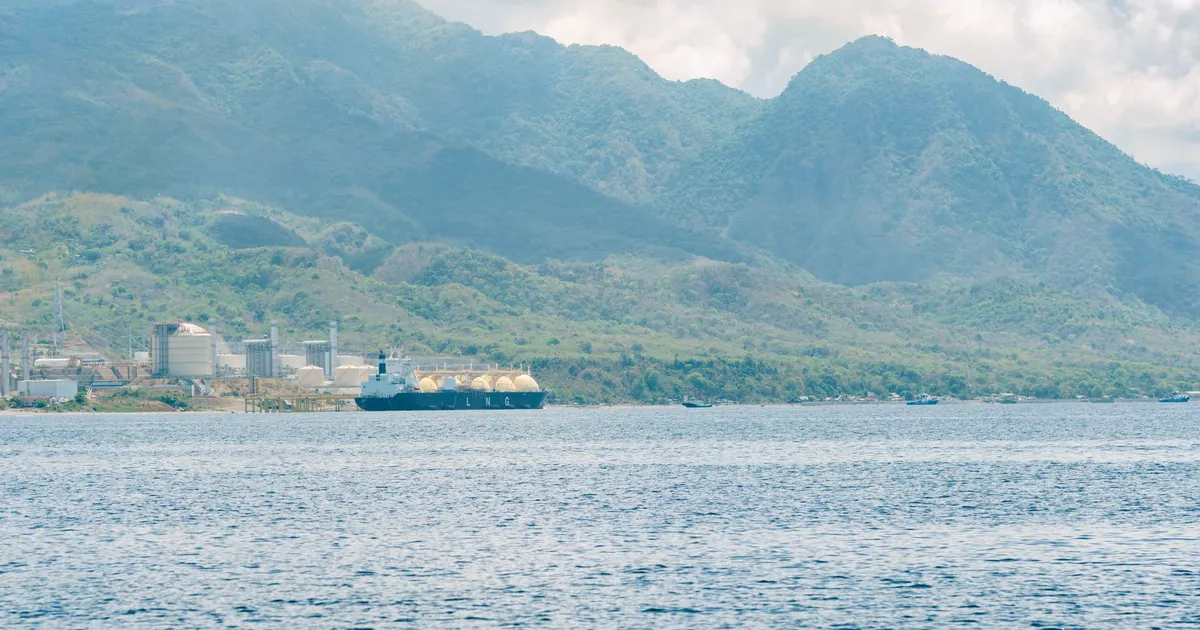 LNG tanker at an industrial port with lush mountains in the background under a cloudy sky.