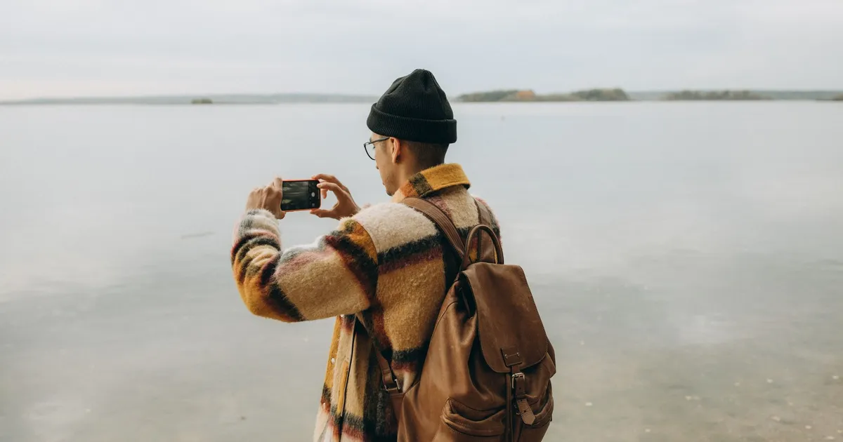 Back view of a man capturing a lake with a smartphone, in a peaceful outdoor setting.