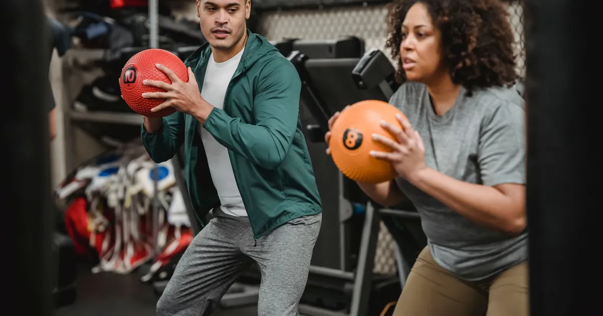 Serious young ethnic male instructor in sportswear looking at plus size African American female doing squat exercise with medicine ball in gym