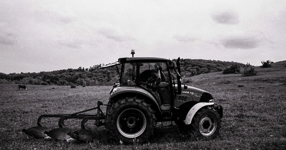 A tractor plows a field in an open countryside, set against a hilly backdrop.