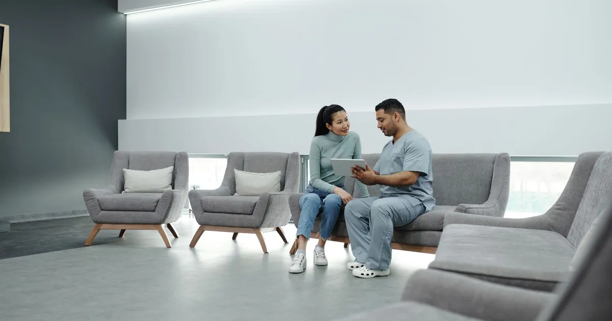 A doctor in scrubs consulting a female patient in a modern clinic waiting room, using a tablet.