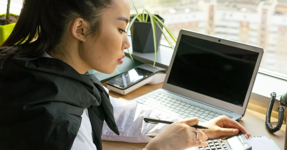 Asian woman working in a bright office, calculating finance on a desk with a laptop.