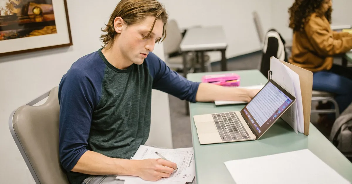 College student writing notes and using a laptop at a classroom desk for exam preparation.