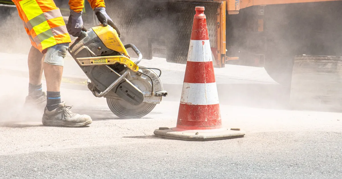 A construction worker cutting asphalt with a large saw next to a traffic cone on a street.