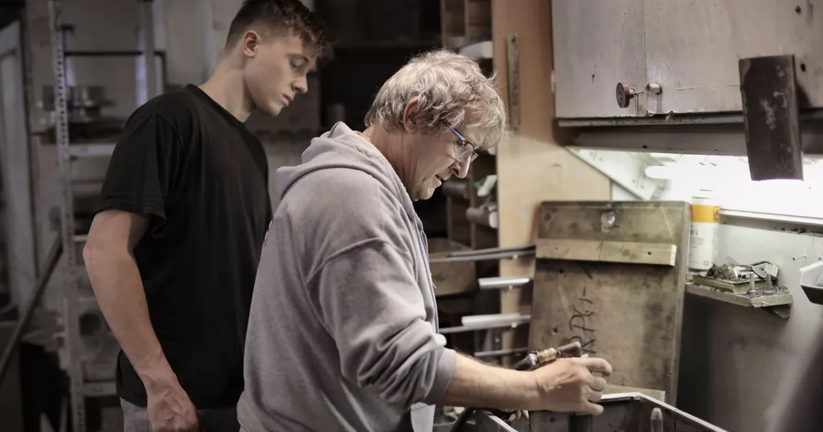 Side view of senior worker in eyeglasses and trainee handling detail on workbench in workshop