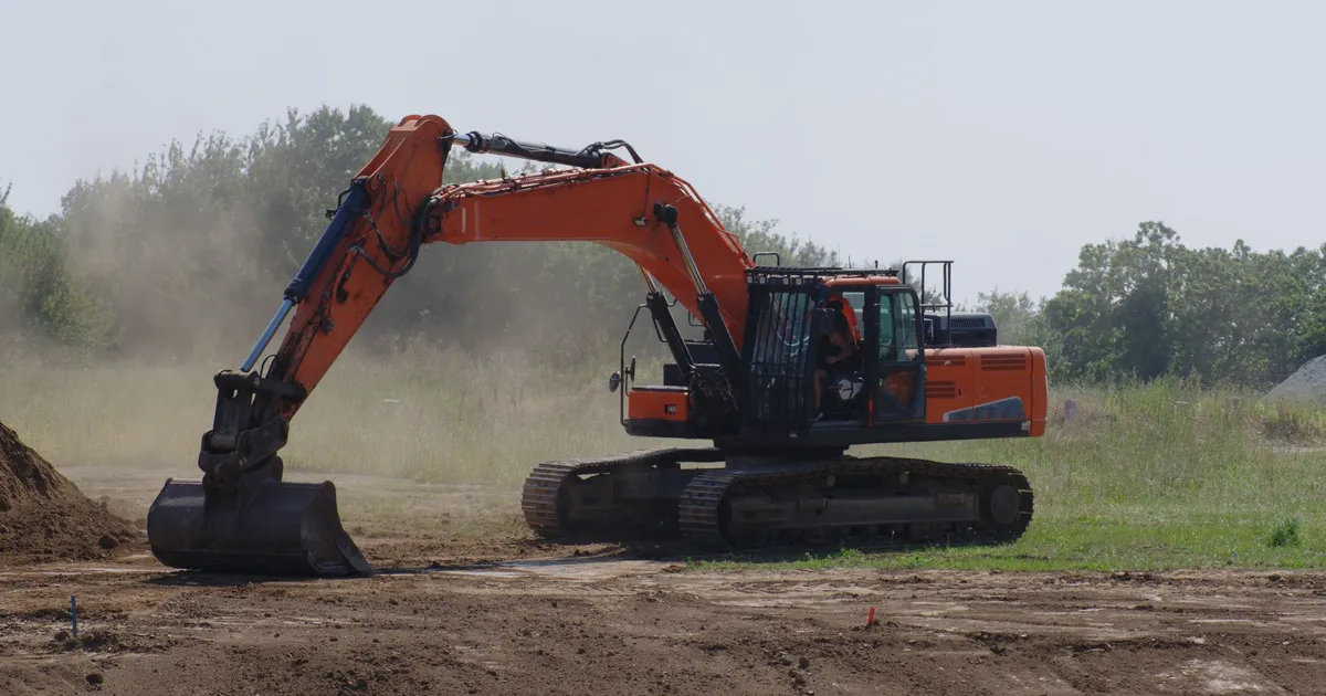 An excavator levels dirt at an outdoor construction site with dust and trees visible.