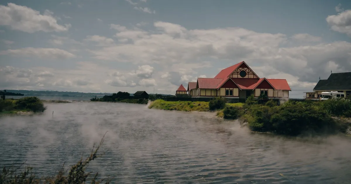 Traditional Maori building by Rotorua Lake with geothermal steam rising, captured in daylight.