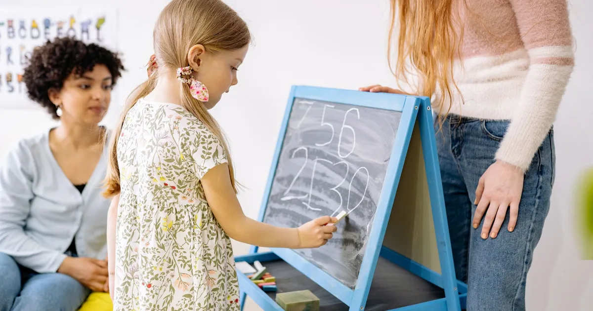 A young girl writes numbers on a chalkboard in a classroom, supervised by adults.