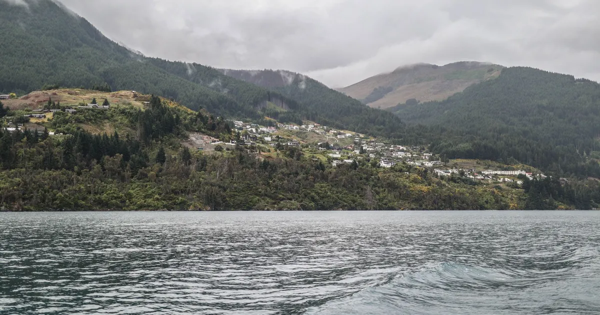 Dramatic landscape of Queenstown's lake surrounded by lush green mountains under a cloudy sky.