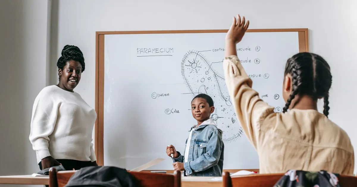 Ethnic girl raising hand while African American female teacher standing near whiteboard with teen boy and explaining task
