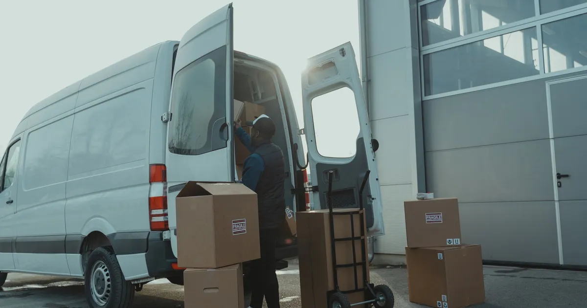 Courier loading cardboard boxes into a delivery van outside a warehouse.