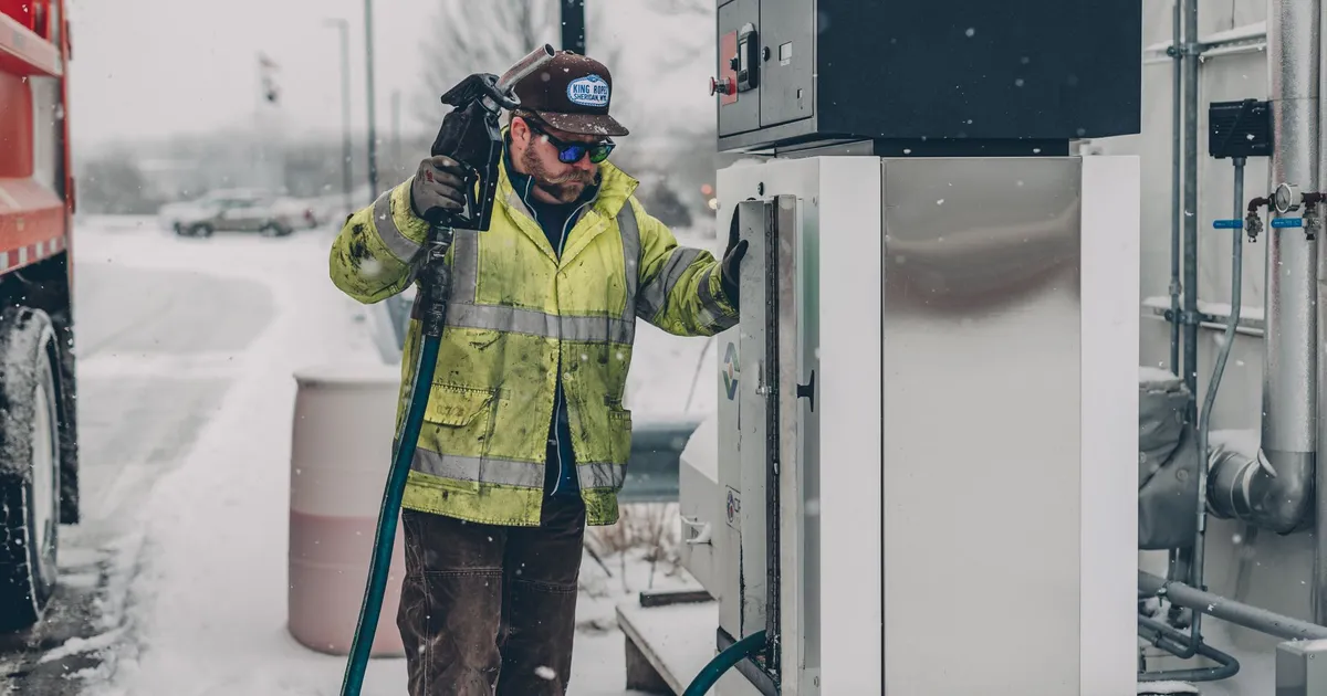 A man wearing a jacket refuels a truck at a gas station during winter with snow falling.
