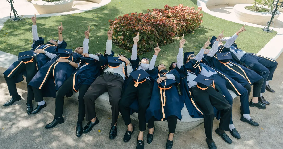 Graduates in caps and gowns celebrate by pointing skyward outdoors on a sunny day.