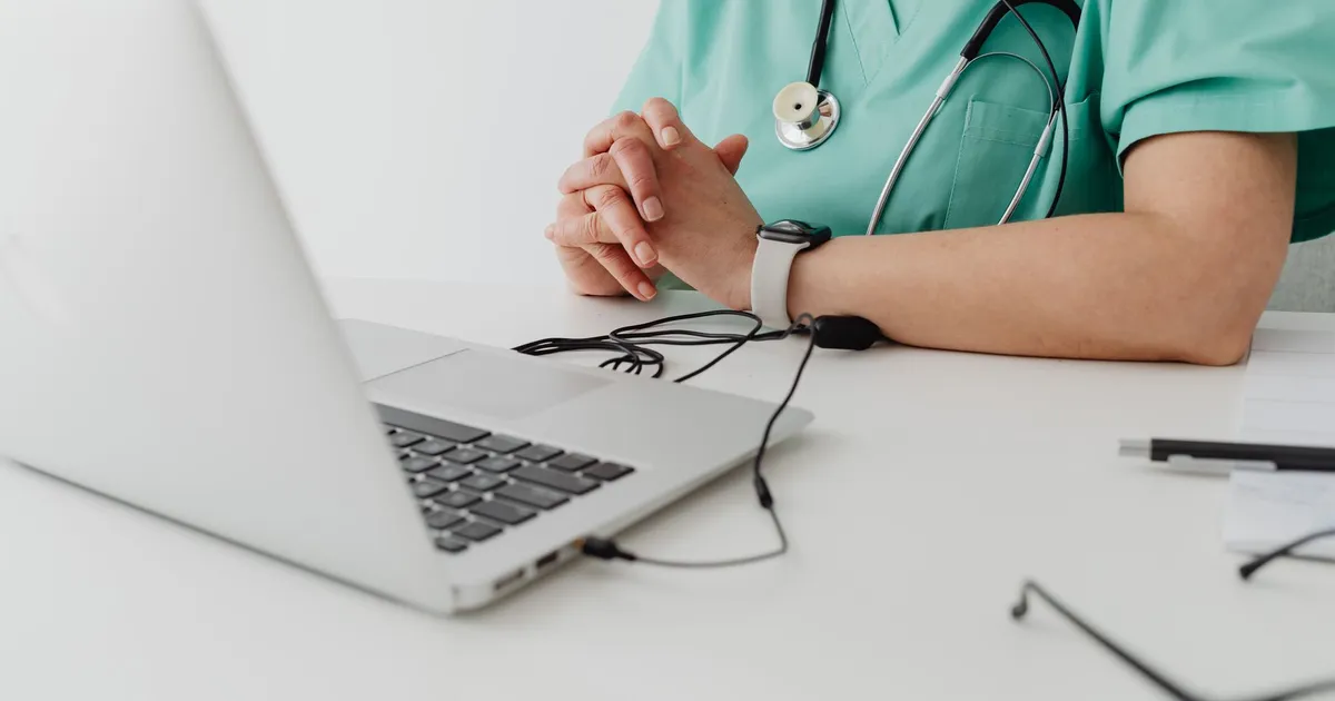 Medical practitioner in scrub suit using a laptop for remote consultation and documentation.