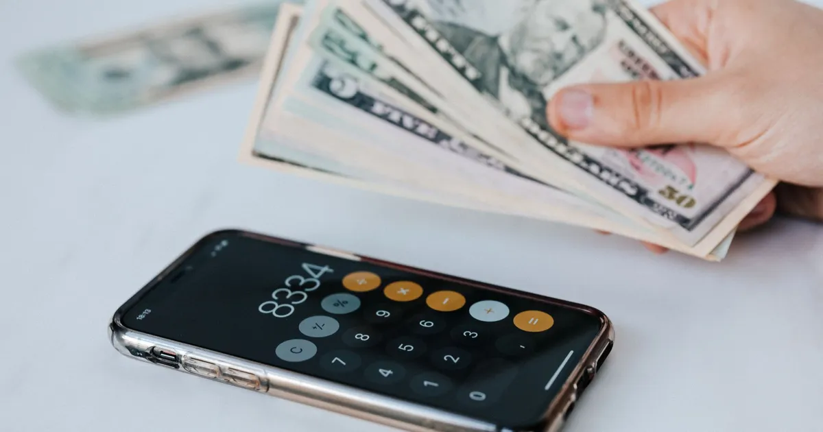 Calculator application on mobile phone screen on white surface near crop anonymous person holding pile of dollars