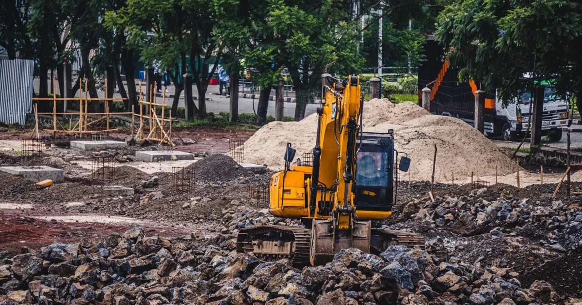 A yellow excavator working at an urban construction site surrounded by rocks and trees.