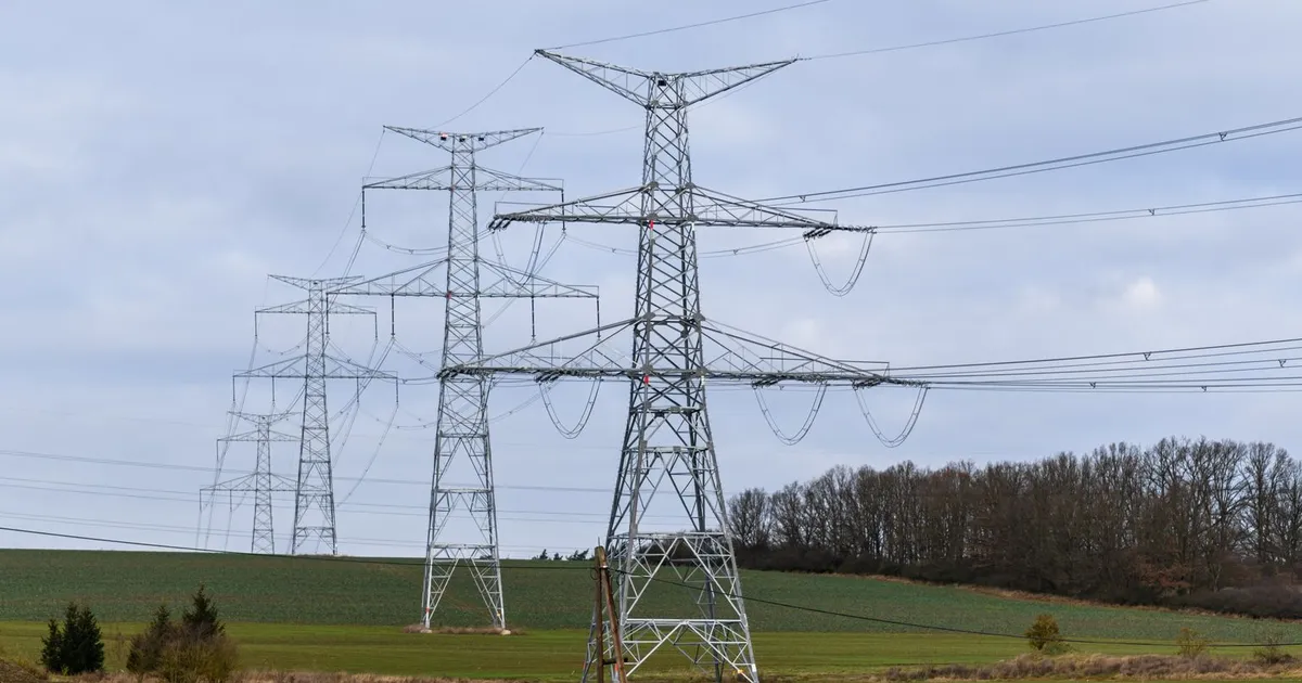 High voltage power lines stretch across a rural field beneath a cloudy sky.