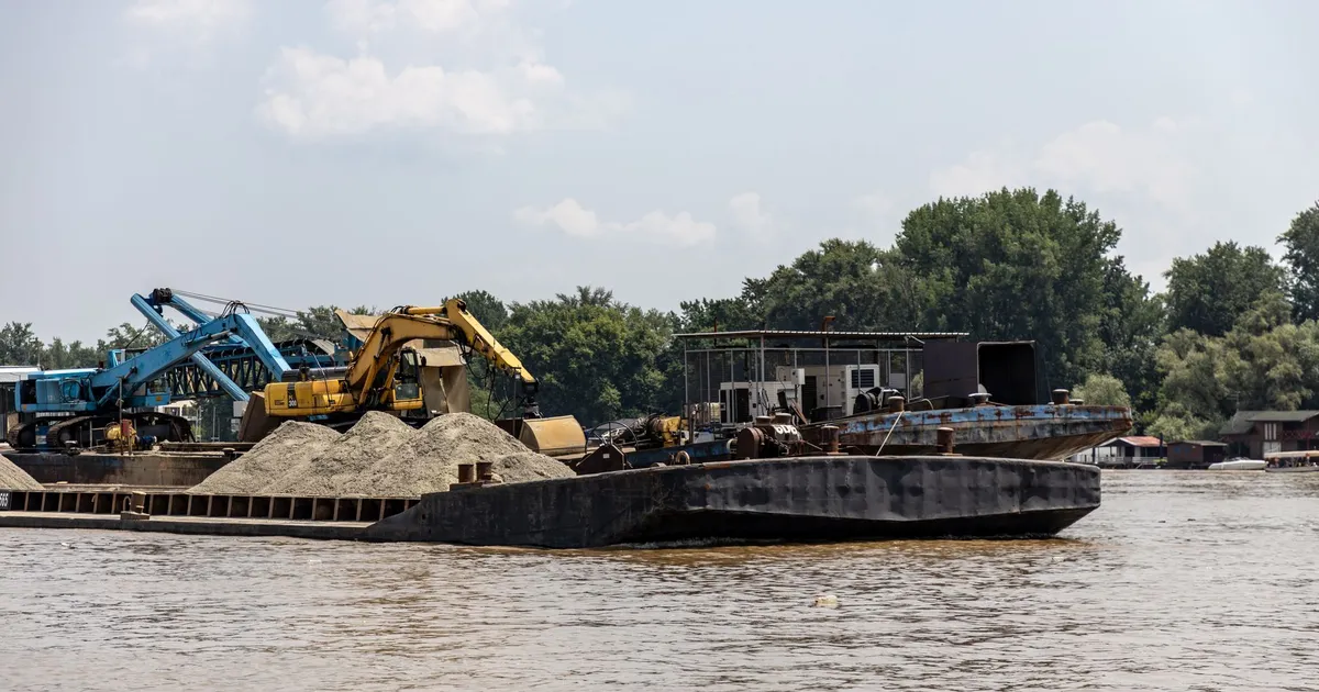 Barge with excavators and sand, working on a river under a bright sky.