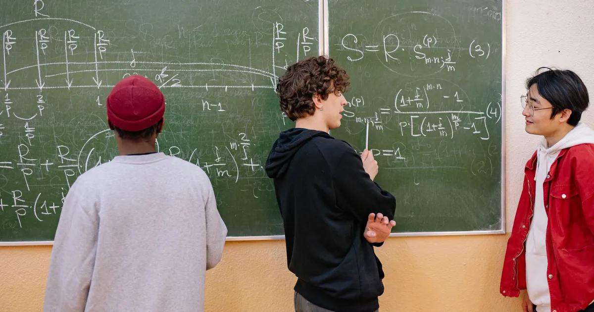 Three students discuss math equations on a classroom blackboard, fostering learning and teamwork.