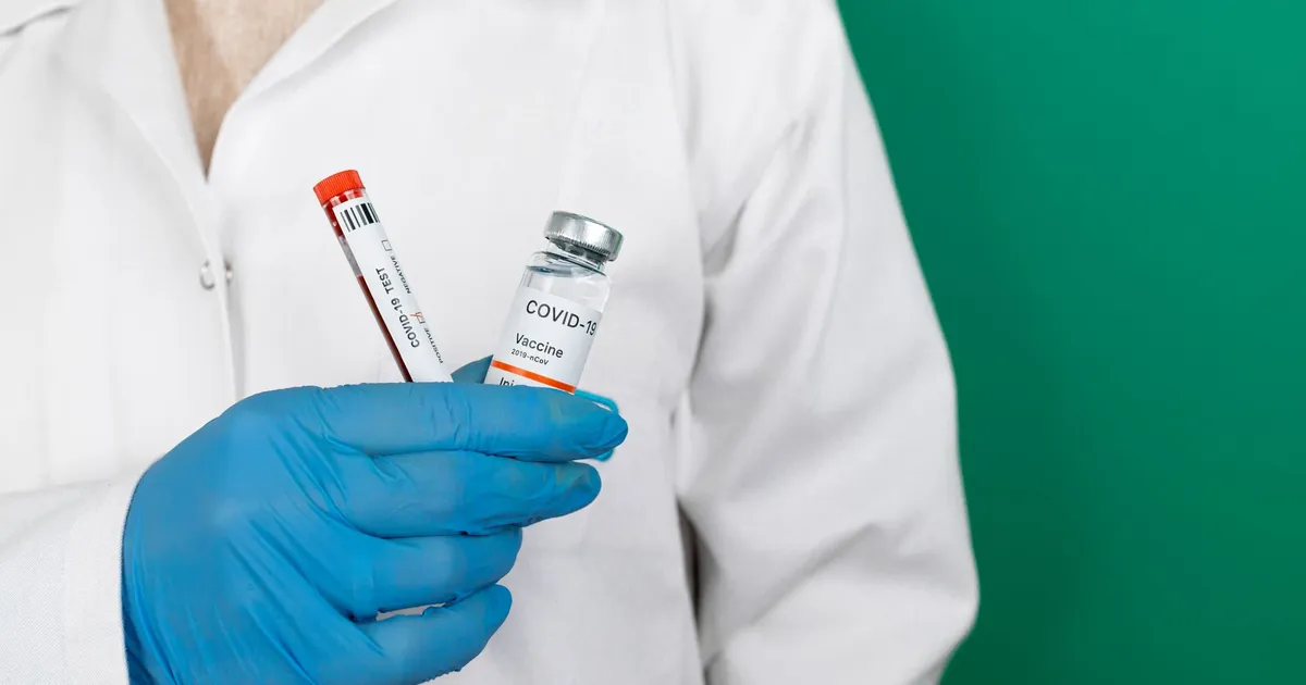 Close-up of a gloved hand holding a COVID-19 vaccine vial and test tube in a lab coat.