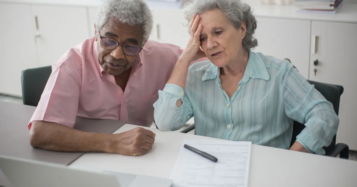 Elderly couple engaged with a laptop, reviewing paperwork at home office desk.