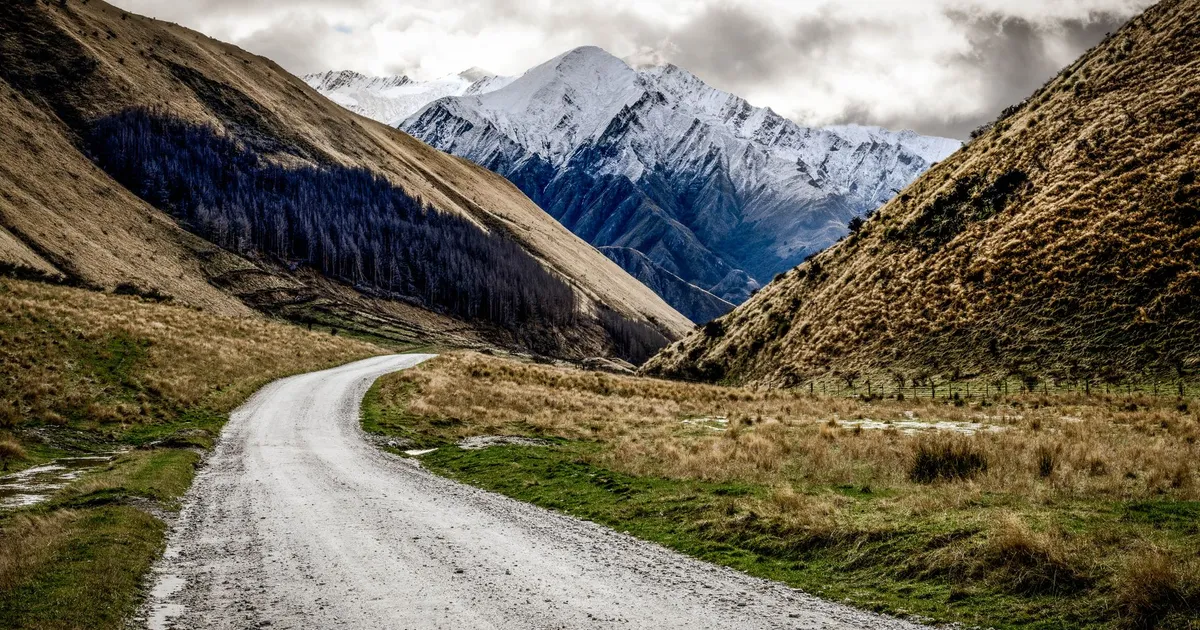 A winding gravel road through New Zealand's picturesque mountains under a cloudy sky.