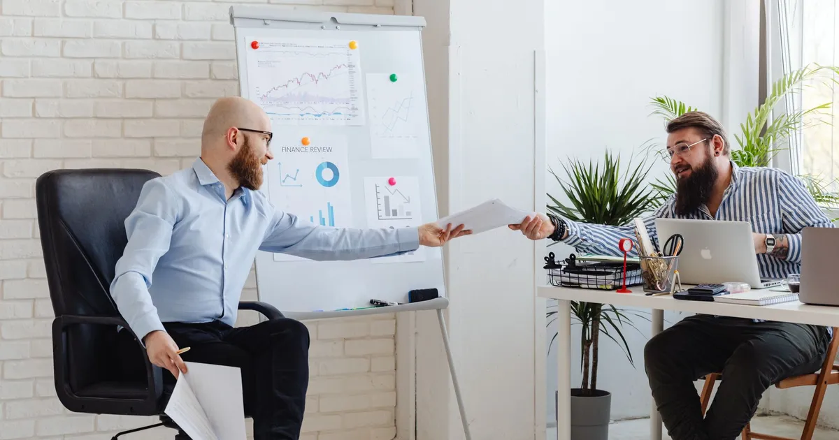 Two business professionals exchanging documents in a modern office setting with charts and graphs.