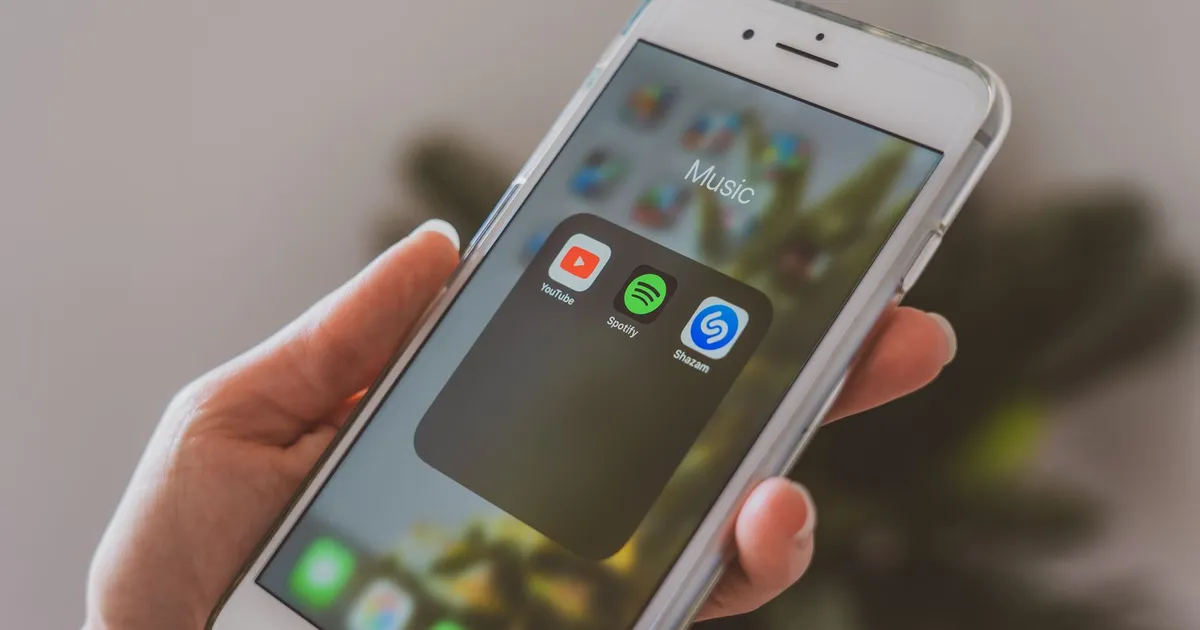 Hand holding a smartphone displaying popular music apps like Spotify and Shazam in a close-up shot.