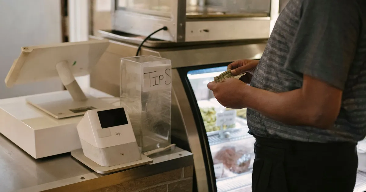 Unrecognizable male in casual clothes standing with money near cash register in grocery store while making purchase near glass showcase