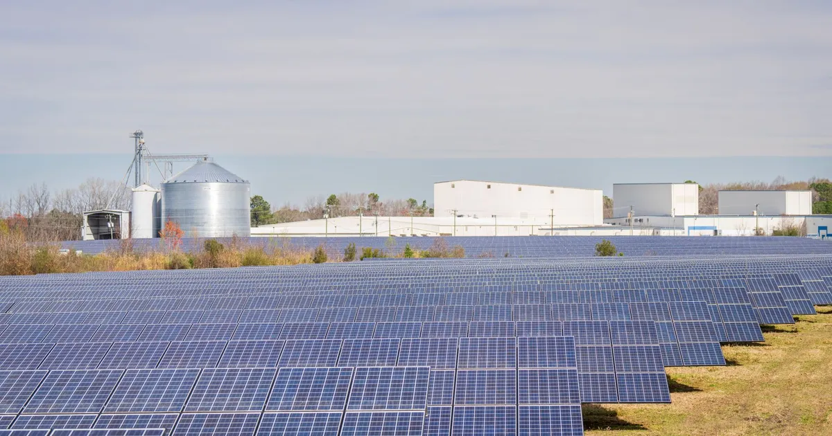 A large solar panel field with warehouses and silos in the background under a clear sky.