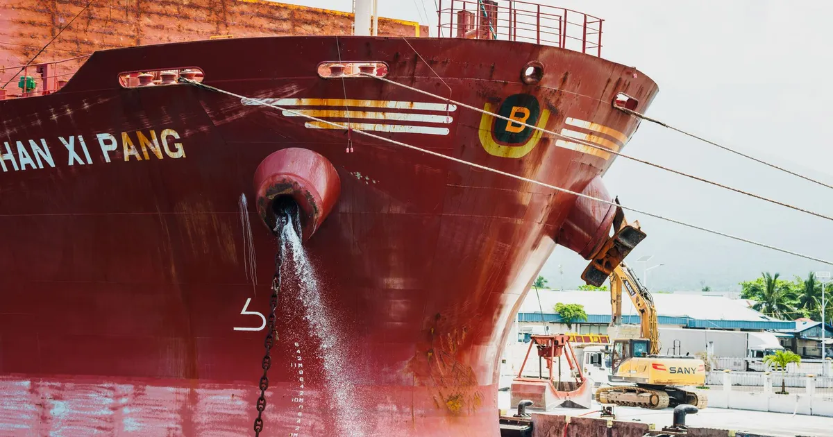 Front view of a large red cargo ship docked, releasing water, with construction equipment nearby.