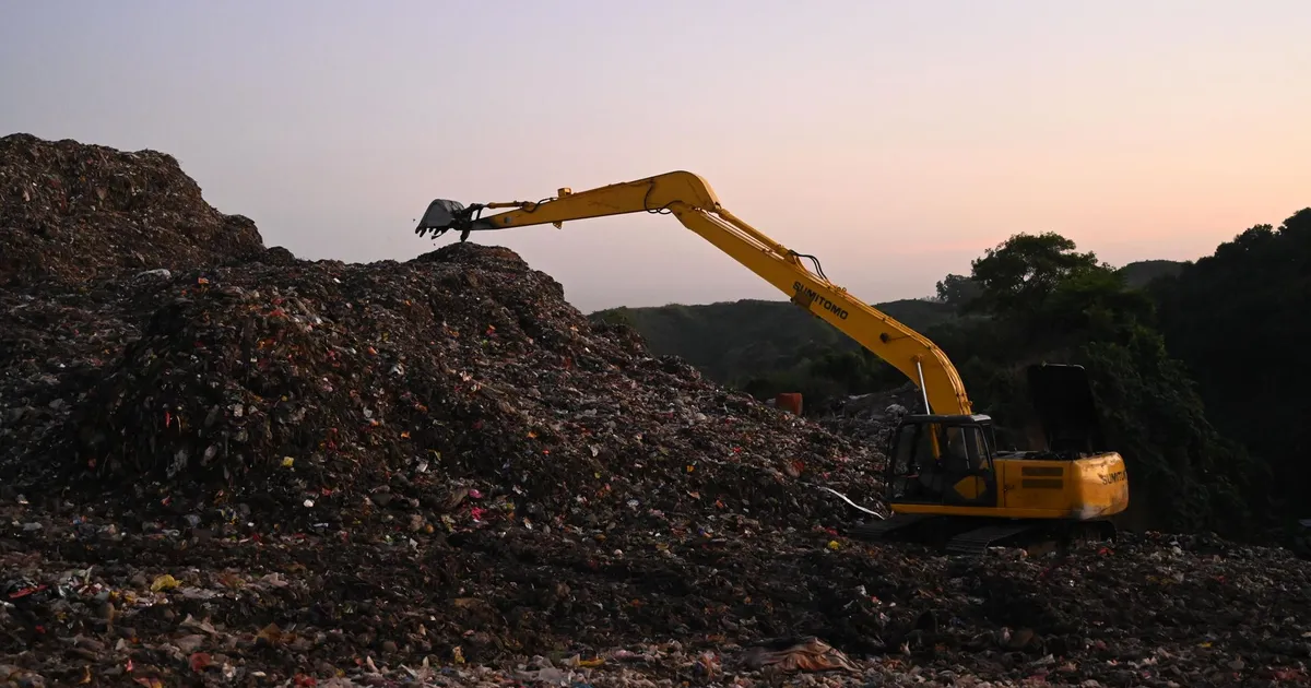 Yellow excavator operates amidst piles of waste in a Bangladesh landfill at sunset.