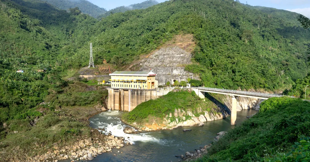 Modern hydroelectric power station with construction located near rippling river and bridge amidst mountainous area covered with green plants in nature on summer day
